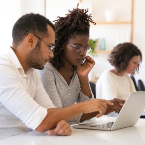 Instructor explaining corporate software specific to intern. Man and woman in casual sitting at desk in classroom, working on laptop, pointing at screen, talking. Education concept